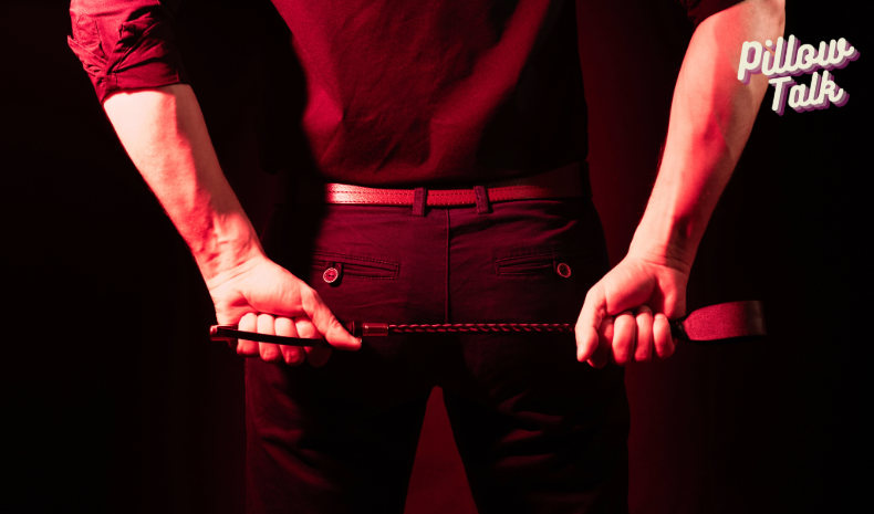 A man stands with his back to us in a sultry red light with a riding crop in both hands across his back