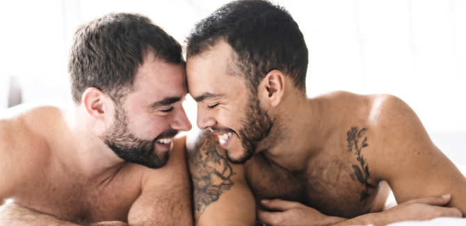 Two men, laying down in bed, shirtless, facing each other with their foreheads pressed against each other. They are smiling and intimate.