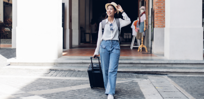 Young person of asian decent walking on the street at an intersection with a rolling suitcase in right hand, loose/breezy, clothing, a beach hat and sunglasses. Woman is smiling and looking around.