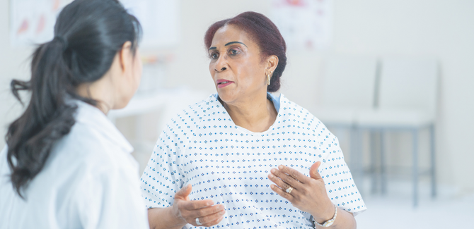 Older Black woman (right) is in hospital gown speaking with doctor (left) who has long black hair in a pony tail, a white lab coat, and is facing the patient (back is at camera). Black woman is animated with her hands with a neutral look on her face.