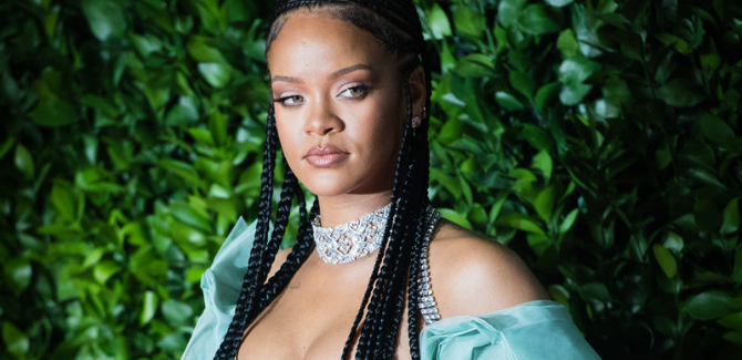 Rhianna stadning in front of greenery, looking to her left, wearing green/sage dress, and a diamond choker necklace.