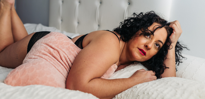 Curvy woman laying on bed on her side wearing a black bra and panties. Woman has black curly hair and is clutching a pink pillow under right arm. Face is neutral and eyes are looking upward.