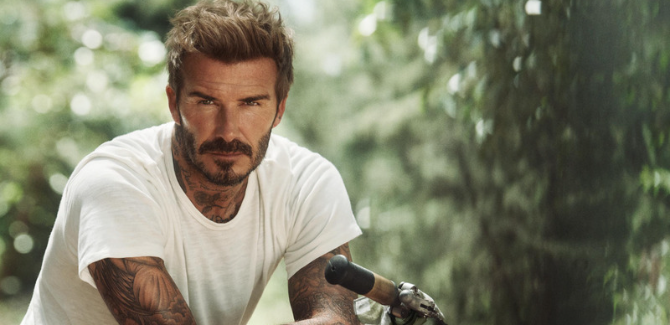 David Beckham stares ahead in a white shirt, siting on a motorcycle, in a forested area.