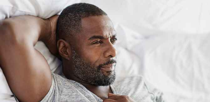 Idris Elba lays on a bed with white sheets, right hand behind his head, staring away from the camera. He is in a gray t-shirt and right hand is cletching his chest.