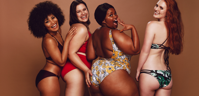 Four women of mixed races and diverse body sizes are stannding in swimwear, rear facing, in front of dark brown backdrop. Womne are posing with their butts arched and emphasized.
