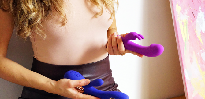 Woman standing by a wall with blonde hair (face not shown) wearing a tight peach bodysuit and leather skirt. She is holding a Bright magenta Rabbit Vibrator in her left hand and a royal blue Rabbit vibrator in her right hand.