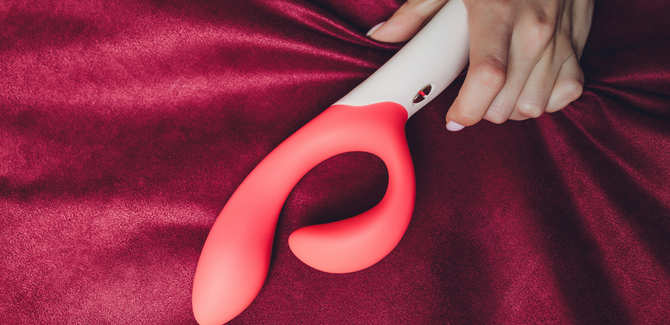 Coral and white rabbit vibrator laying on a red cloth and a white hand with white painted nails is grasping it.