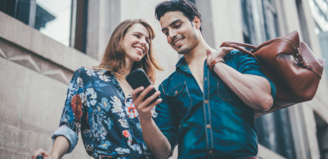 Young man and woman on the sidewalk, with travel bags. Man (RIGHT) has brown leather bag thrown over left shoulder. He is smiling and looking at his phone. Woman (LEFT) has beige suitcase, a floral top is looking at man, smiling.