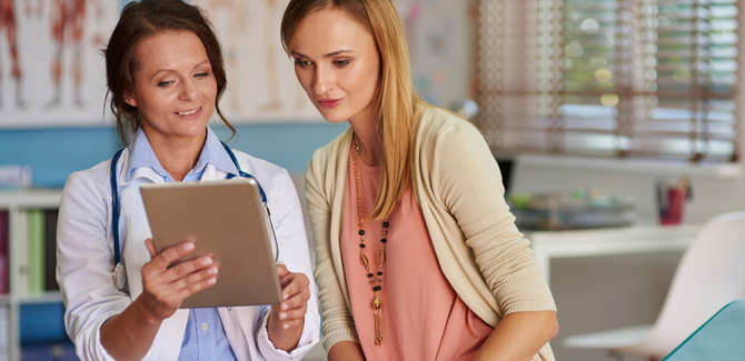Woman (right) in peach top and beige sweater is consulting with a doctor (LEFT). Both are looking at a tablet the doctor is holding.