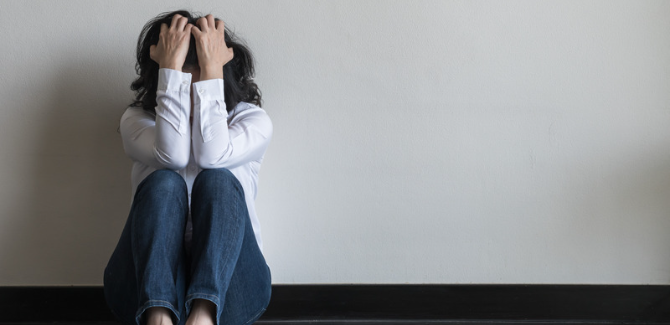 person sitting on the floor with arms in front of face and hands clutching brown hair in distress, wearing jeans and white long sleeve shirt.