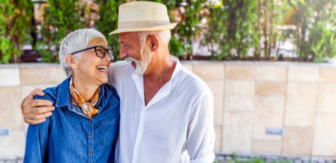 Older man and woman, standing together, smiling at each other outside. Man (RIGHT) is wearing loose, white shirt and fedora hat, his right arm around woman (LEFT) who is wearing loose jean shirt and orange neckerchief. 