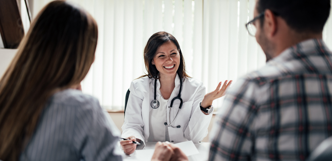 Couple is sitting at a desk, facing a doctor, (their backs are at camera) who is giving them advice. Doctor is woman with white coat and stethoscope around her neck.