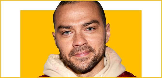 Jesse Williams staring straight ahead, small grin, wearing a beige color hoodie and red jacket. Image is on white background with a yellow square and a yellow frame surrounds image..